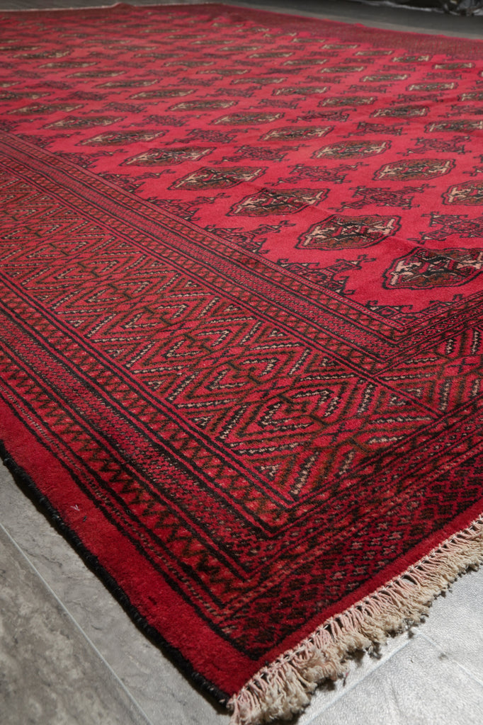 A red patterned rug with dark borders and cream fringe, photographed at an angle on a gray floor.