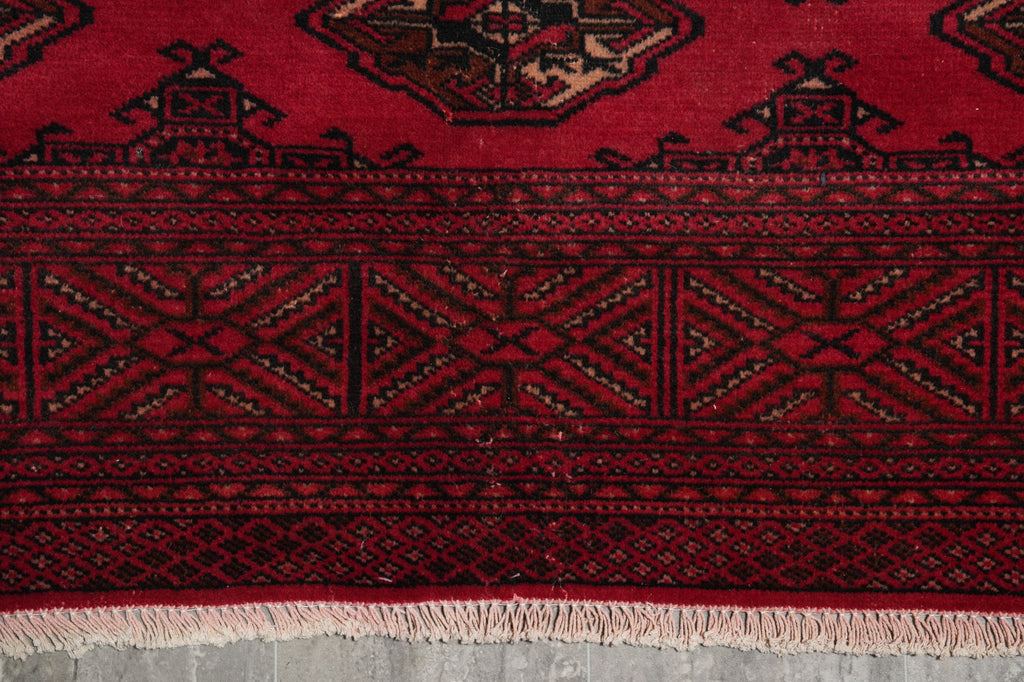 A close-up of a traditional patterned rug with a deep red field, black geometric borders, and light beige fringe at the bottom.