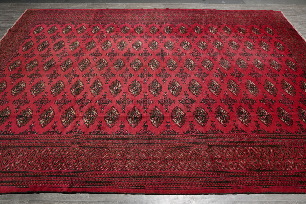 A large red oriental rug with an intricate repeating geometric and floral pattern in dark brown and black, laid out flat on a gray tiled floor.