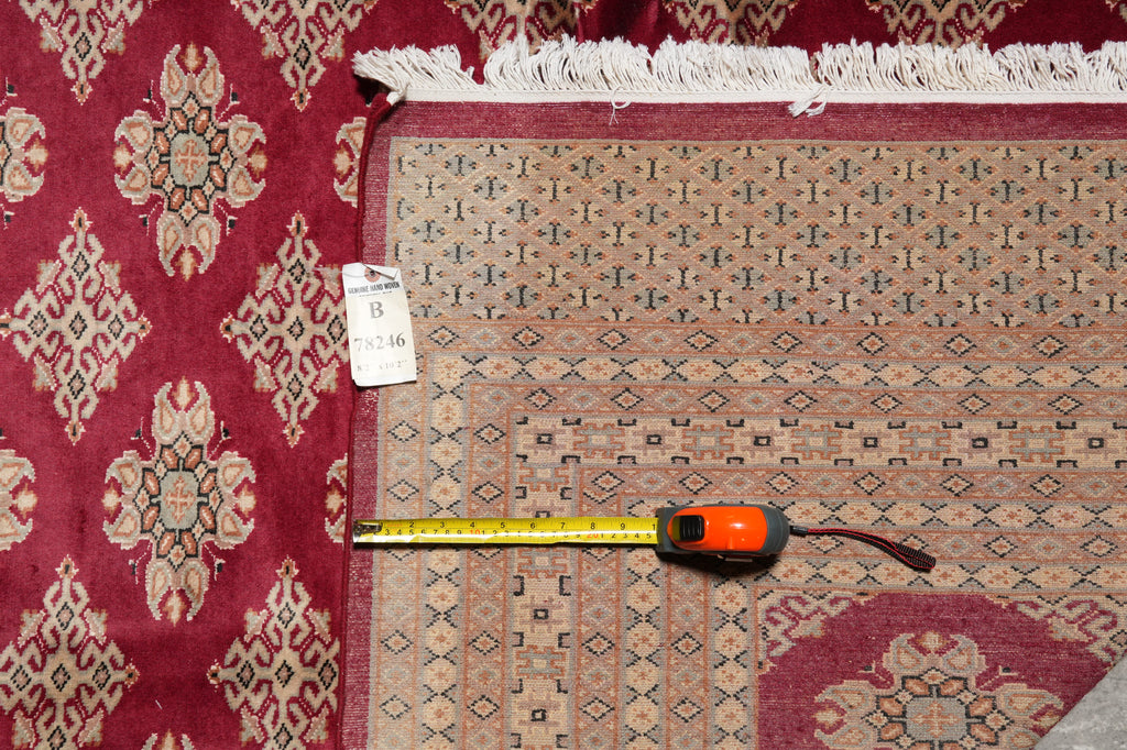 A rectangular area rug with a beige and red geometric pattern and white fringe, shown alongside a matching red patterned textile and a tape measure for scale.