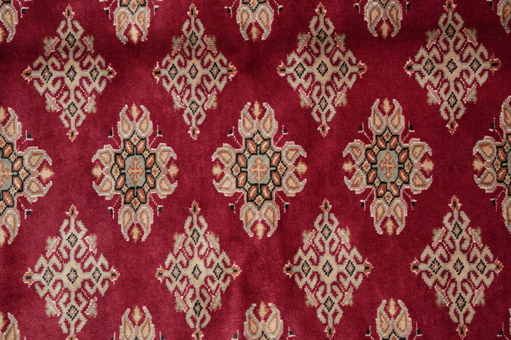 A close-up of a red patterned rug or fabric with repeating ornate medallion motifs in beige, cream, black, and muted blue.