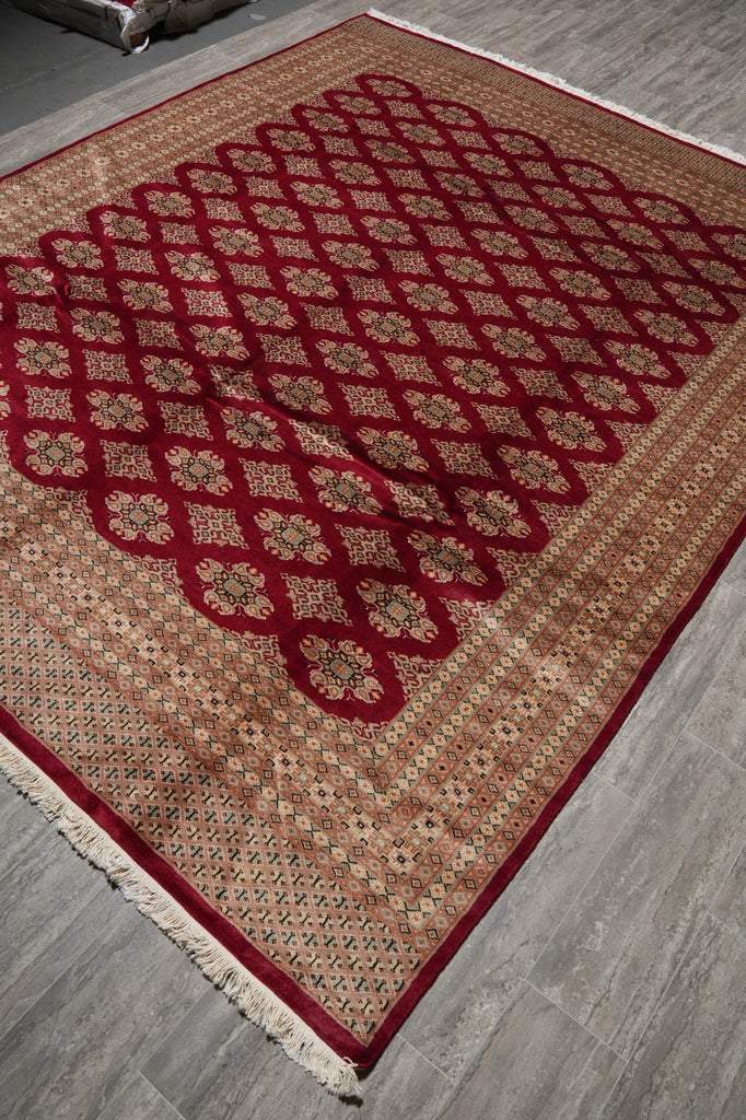 A rectangular area rug with a traditional Persian-style design, featuring a deep red lattice pattern over a beige and brown background, laid out on a gray wood floor.