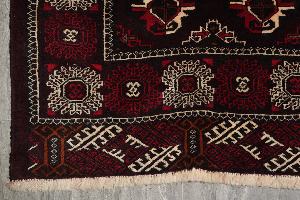 A close-up of a traditional patterned rug corner, showing a dark red and black field with cream and red geometric motifs, a decorative border, and cream fringe.