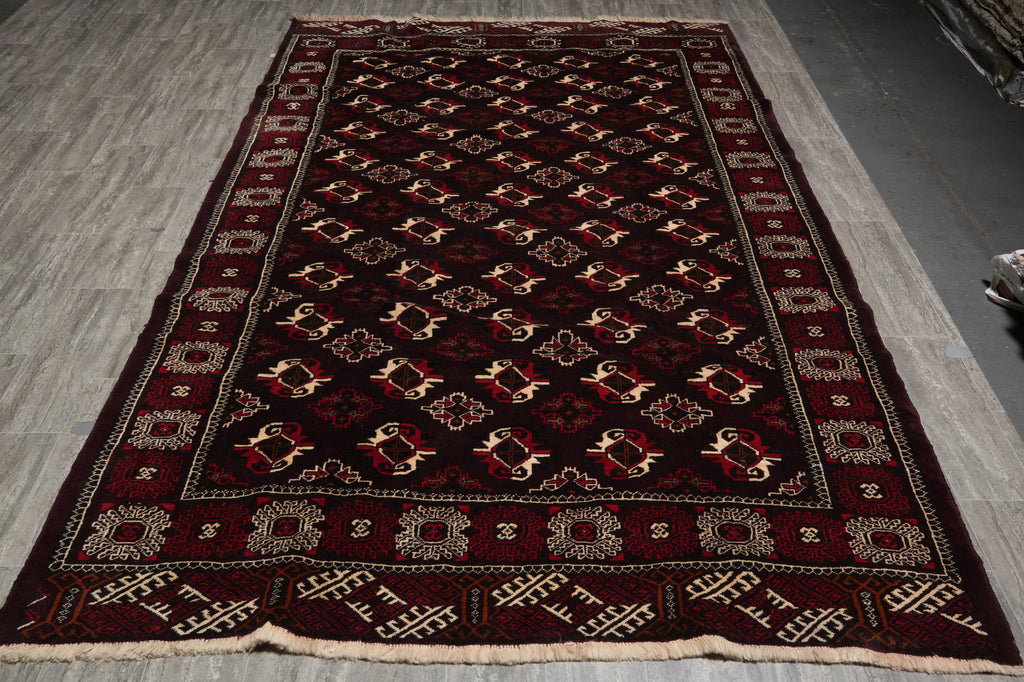 A rectangular dark red and black patterned rug with cream and red motifs, laid flat on a gray wood floor.