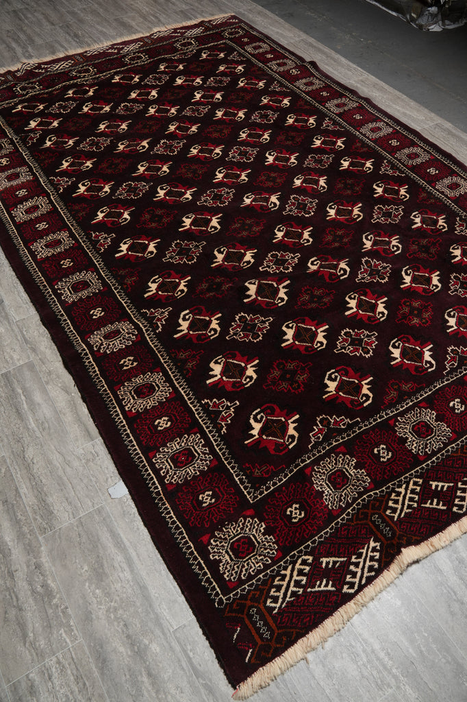 A dark red and black patterned area rug with beige geometric borders and motifs, laid out diagonally on a gray tiled floor.