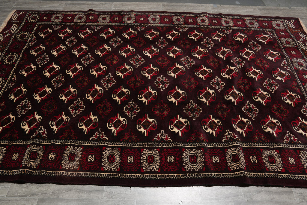 A large rectangular area rug laid flat on a gray tiled floor, featuring a dark red and black background with repeating cream and red geometric motifs and ornate borders.