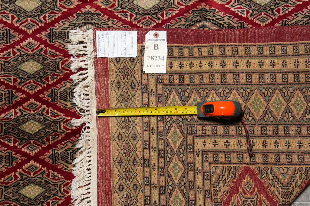 A patterned area rug with cream fringe is shown laid flat on a matching patterned surface, with a yellow tape measure and price tag visible along the left edge to indicate size and condition.