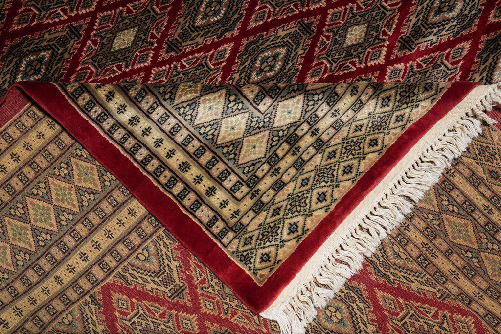 A close-up of a patterned area rug showing its corner, with cream fringe, a deep red border, and a beige field with black geometric motifs over a red background.