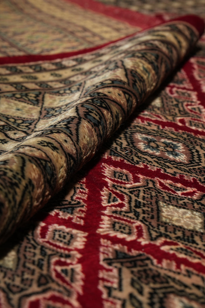 A close-up of a patterned area rug with one corner rolled back, showing detailed woven motifs in red, beige, black, and brown tones.