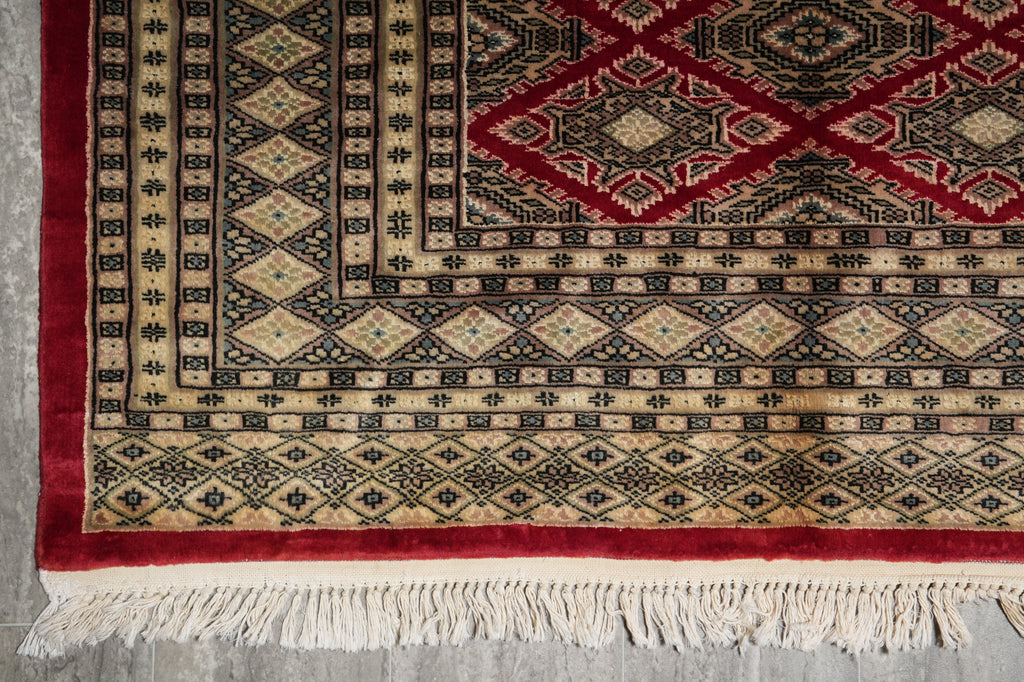 A close-up of a patterned rug corner showing cream, red, and black geometric motifs with white fringe tassels on a gray floor.