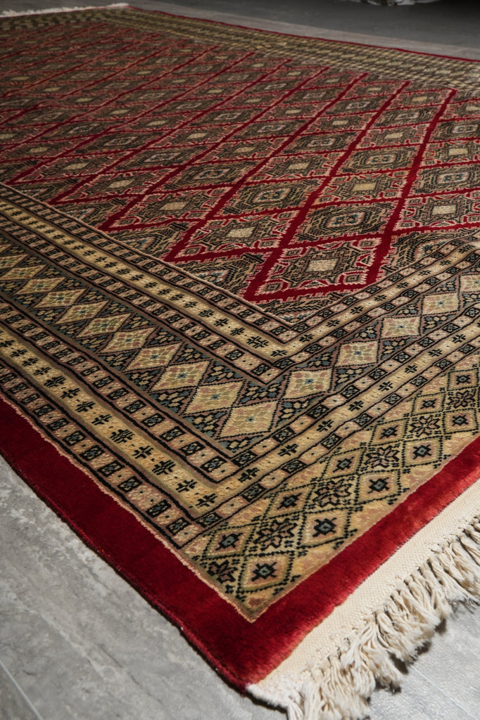 A traditional patterned rug with red borders, beige and black geometric motifs, and cream fringe, shown laid out on a gray floor.