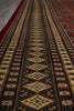 Load image into Gallery viewer, A close-up view of a patterned area rug with geometric diamond motifs in beige, brown, black, and muted red tones, shown laid out on a floor.