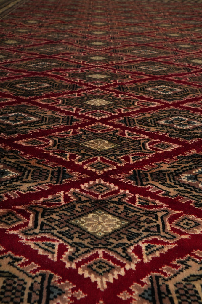 A close-up of a woven rug or textile with a repeating geometric diamond pattern in deep red, black, beige, and brown tones.