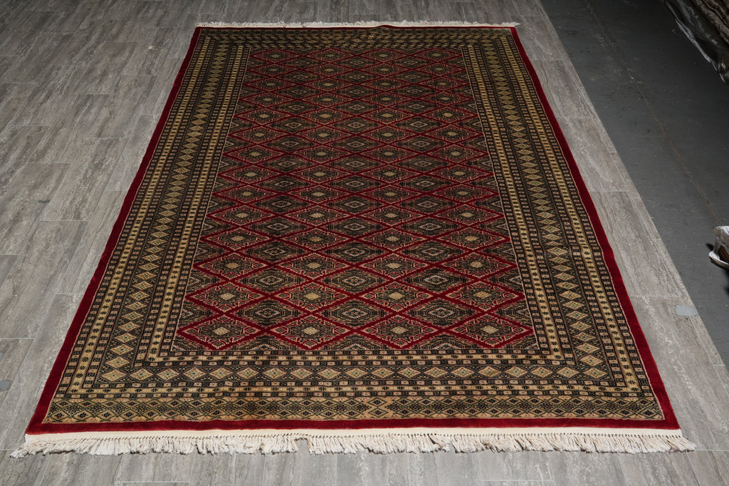 A rectangular area rug with a traditional geometric pattern, featuring a deep red central field, a dark brown border, and cream accents, laid out on a gray wood floor.