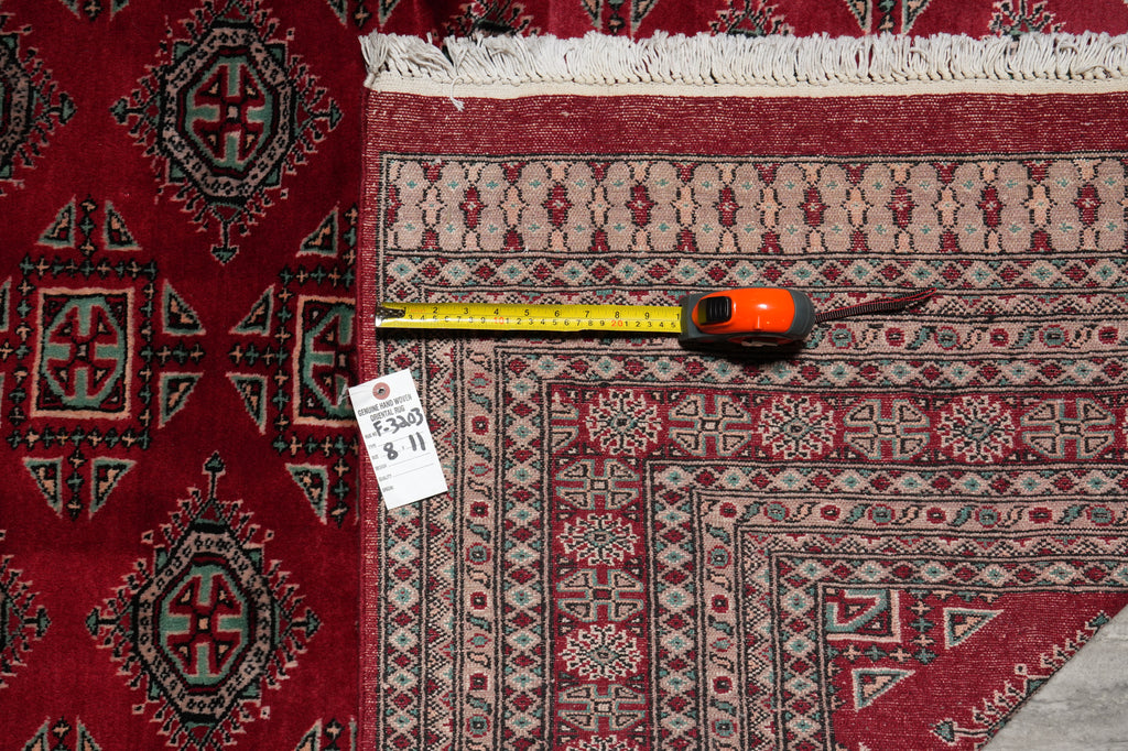 A close-up of a patterned area rug with a red border and cream fringe, shown alongside a yellow tape measure and a white size tag on a gray floor.