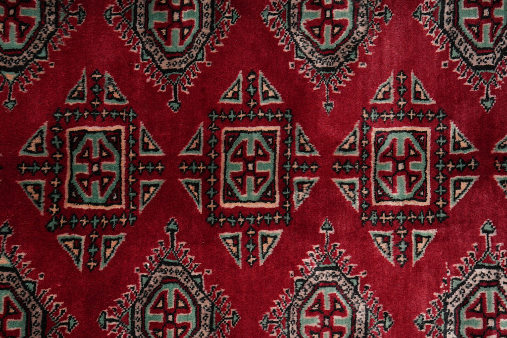 A close-up of a red patterned rug with repeating geometric medallion motifs in black, beige, and muted blue-green.