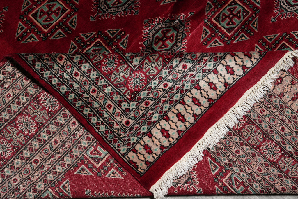 A close-up of a traditional patterned rug with deep red, beige, black, and gray geometric motifs and white fringe.