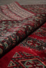 Load image into Gallery viewer, A close-up of a folded patterned rug showing a deep red field with intricate black, beige, and teal geometric and floral motifs.