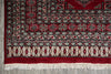 Load image into Gallery viewer, A close-up of a traditional patterned rug corner showing a deep red field, cream and black geometric borders, and white fringe tassels on a gray floor.