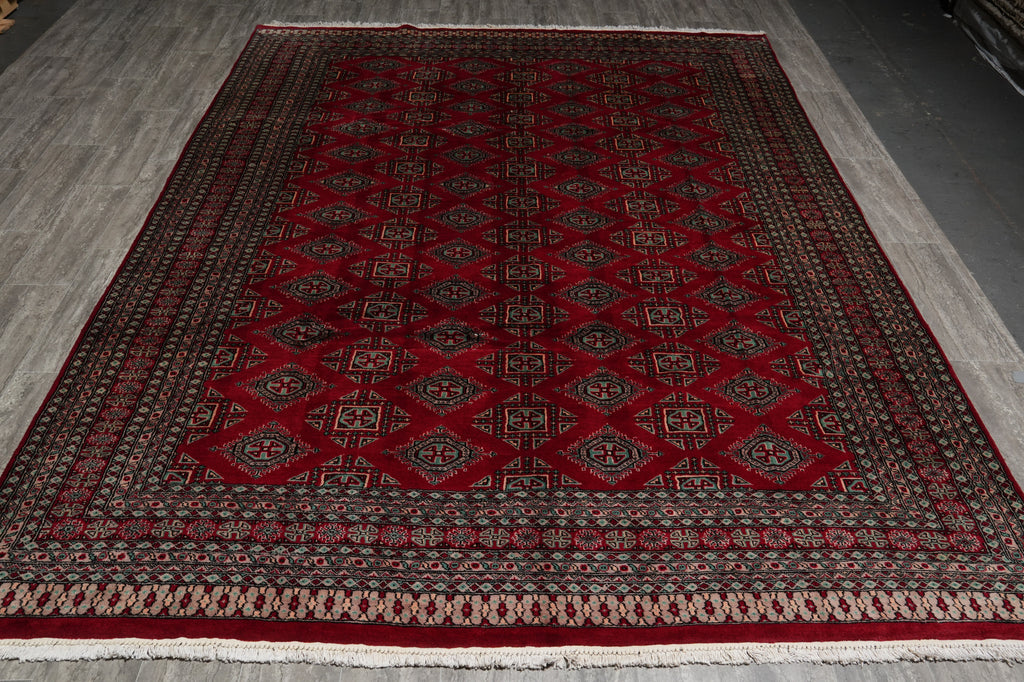 A rectangular red oriental rug with a traditional geometric pattern and fringed edges, laid out on a gray wood floor.