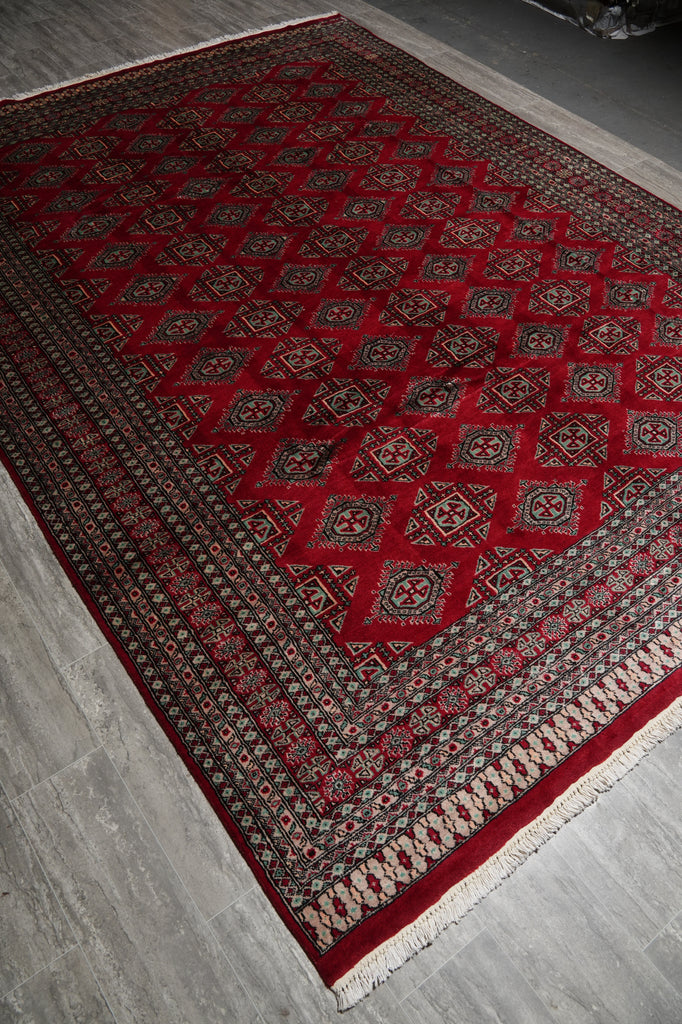 A traditional Persian-style area rug with a deep red field, intricate geometric and diamond patterns in black, beige, and white, and a matching red border, shown laid out on a gray tiled floor.