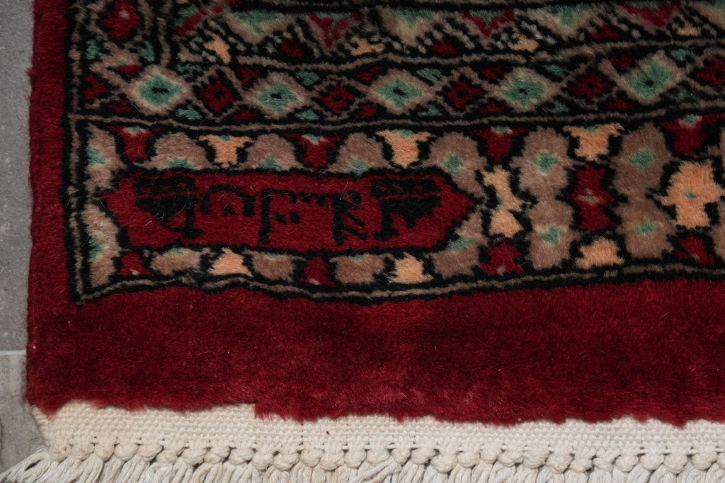 Close-up of a patterned rug corner showing a deep red field, a dark border with beige and teal motifs, and a cream fringed edge.