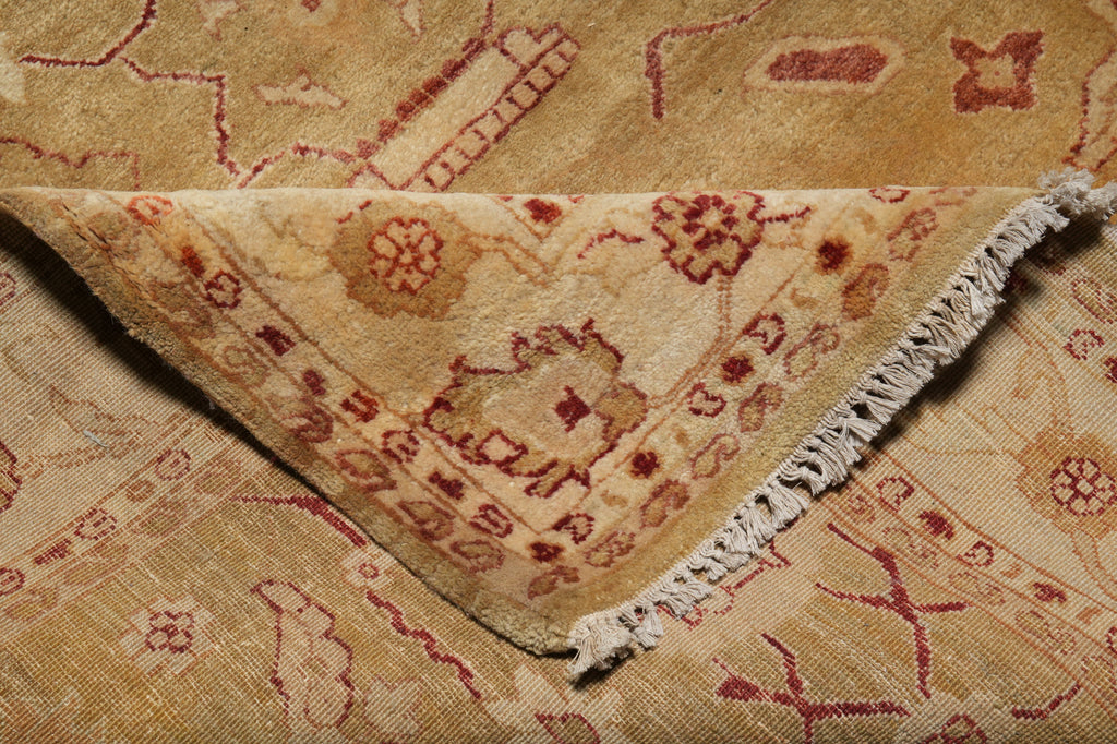 A close-up of a patterned area rug with a folded corner showing the fringed edge. The rug has a warm beige and tan base with dusty red and muted brown geometric and floral motifs.