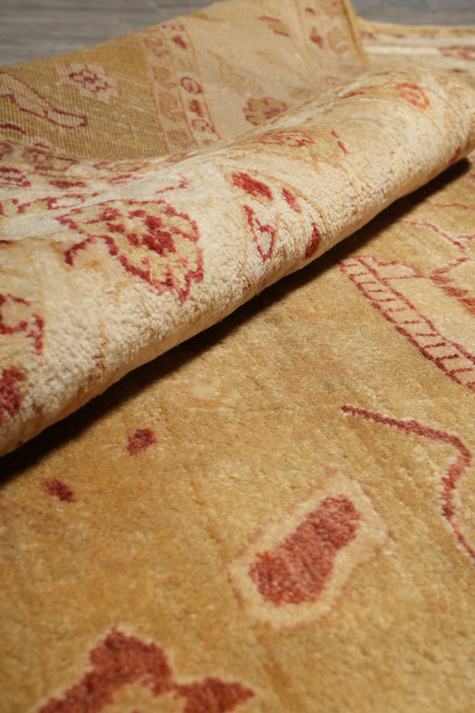 A close-up of a plush area rug with a rolled corner, showcasing its golden-yellow pile and faded red traditional patterns.