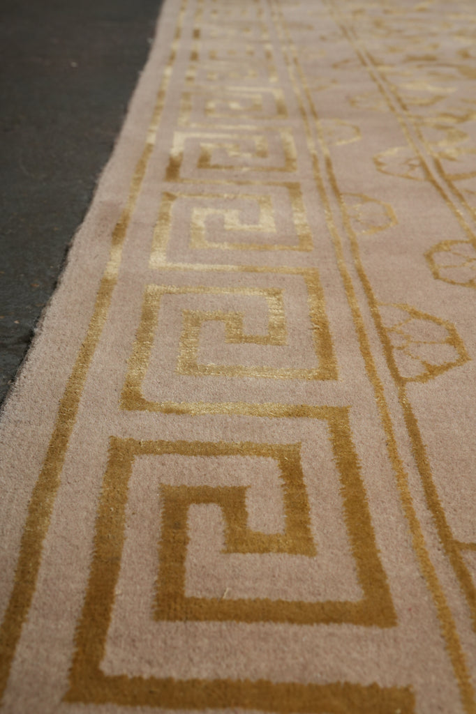 Close-up of a beige textile or rug with a metallic gold Greek key pattern and decorative border details.