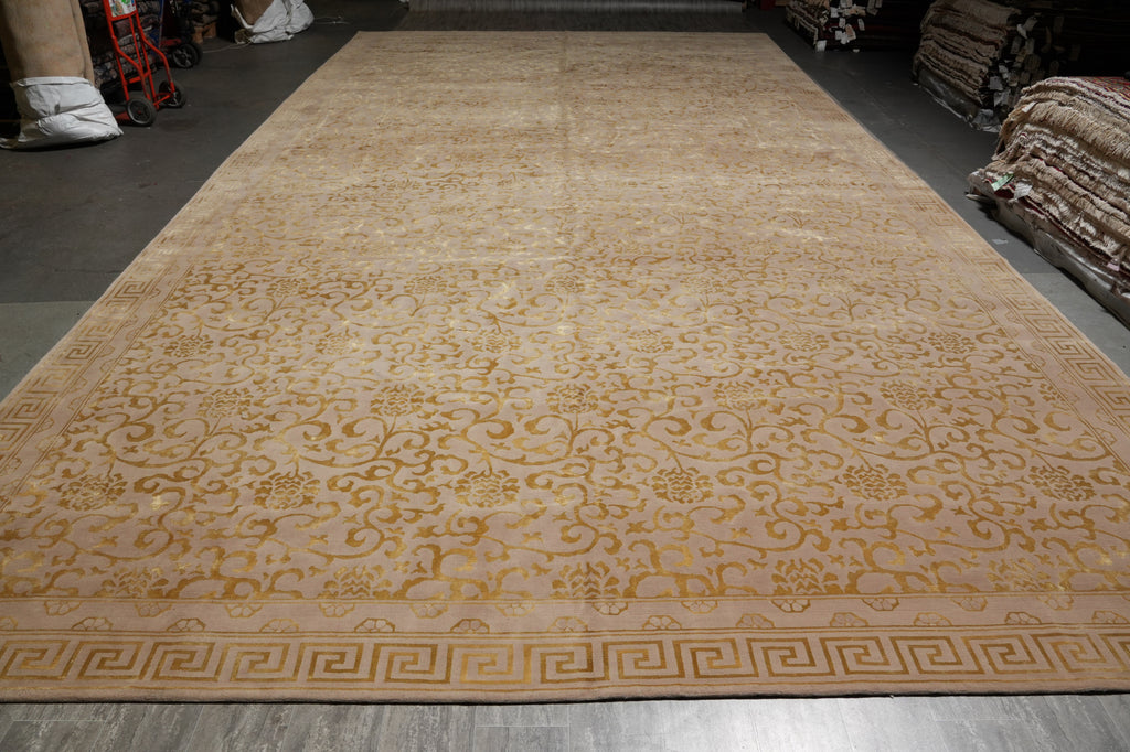 A large rectangular area rug laid out in a warehouse or showroom, featuring a warm beige and tan floral pattern with a Greek key border.