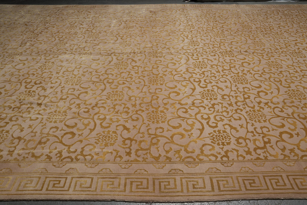 A large rectangular area rug with an intricate golden-brown floral and scroll pattern on a warm beige background, featuring a Greek key border along the bottom edge.