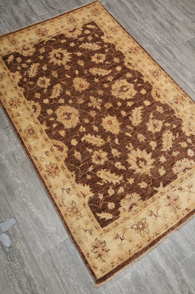 A rectangular area rug with an ornate floral pattern, shown from an overhead angle on a gray wood-look floor. The rug features a dark brown center field with beige floral motifs, surrounded by a wide beige border with smaller floral details.