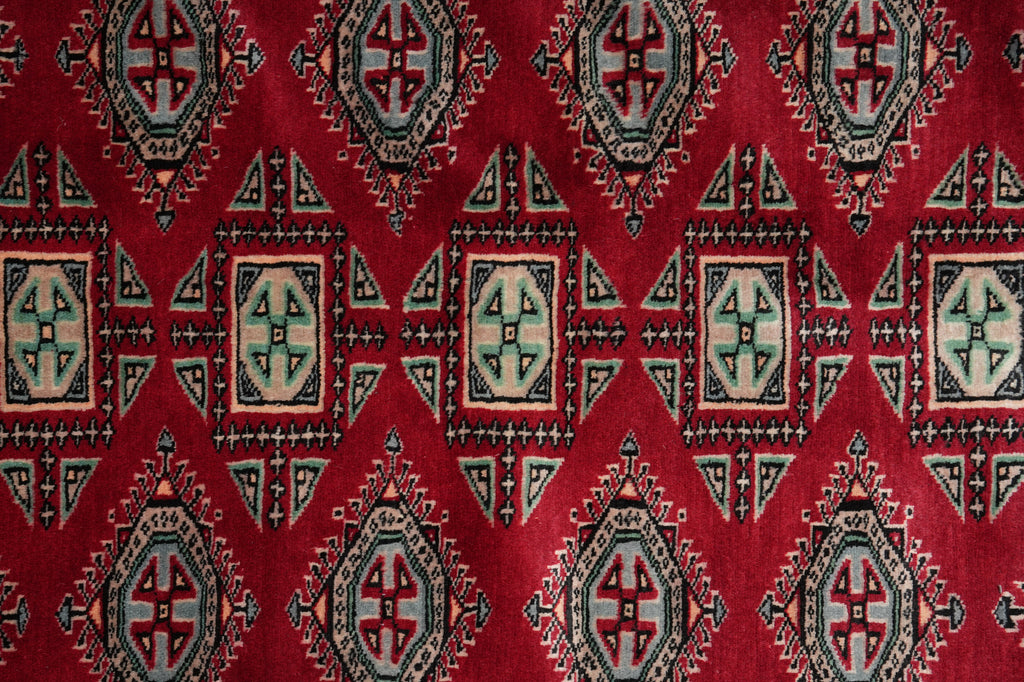 A close-up of a red patterned textile or rug featuring repeating geometric diamond motifs in beige, black, and muted blue-gray.