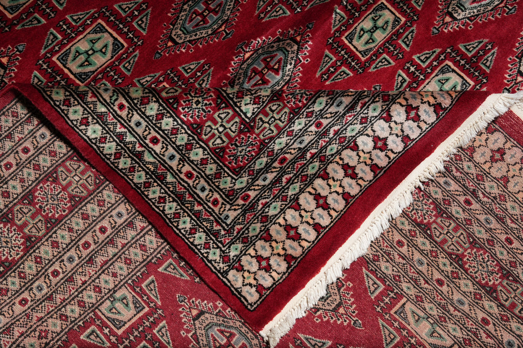 A close-up of a traditional patterned rug with a deep red field, intricate geometric and floral motifs in beige, black, and muted green, and white fringe along one corner.