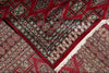 Load image into Gallery viewer, A close-up of a traditional patterned rug with a deep red field, intricate geometric and floral motifs in beige, black, and muted green, and white fringe along one corner.