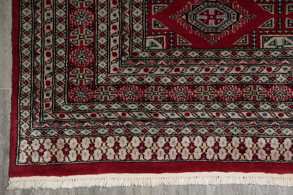 A close-up of a traditional patterned rug corner, showing a deep red field, geometric borders, and cream fringe on a gray floor.