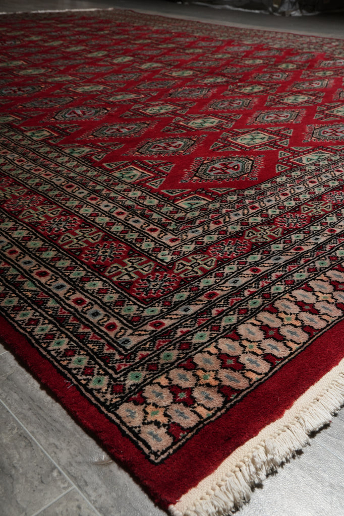 A traditional patterned area rug with a deep red field, cream and black geometric borders, and cream fringe, shown laid out on a gray tiled floor.