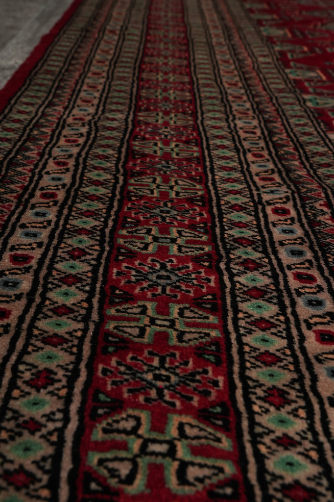 A close-up view of a patterned rug with horizontal bands of red, black, beige, and small turquoise accents in a geometric and floral design.
