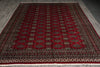 Load image into Gallery viewer, A traditional Persian-style area rug with a deep red field, featuring a repeating diamond pattern in beige, black, and green, bordered by ornate red and beige patterns, laid out on a gray tiled floor.