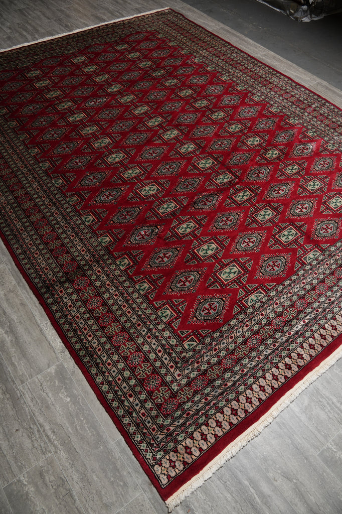 A traditional oriental area rug with a deep red field, featuring a repeating diamond pattern and ornate borders in beige, brown, and black, laid out on a gray tiled floor.
