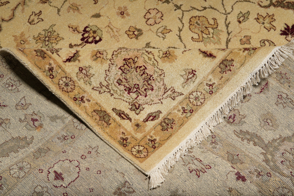 A close-up of a patterned area rug corner with cream and tan tones, floral motifs, and white fringe, laid over another matching rug.