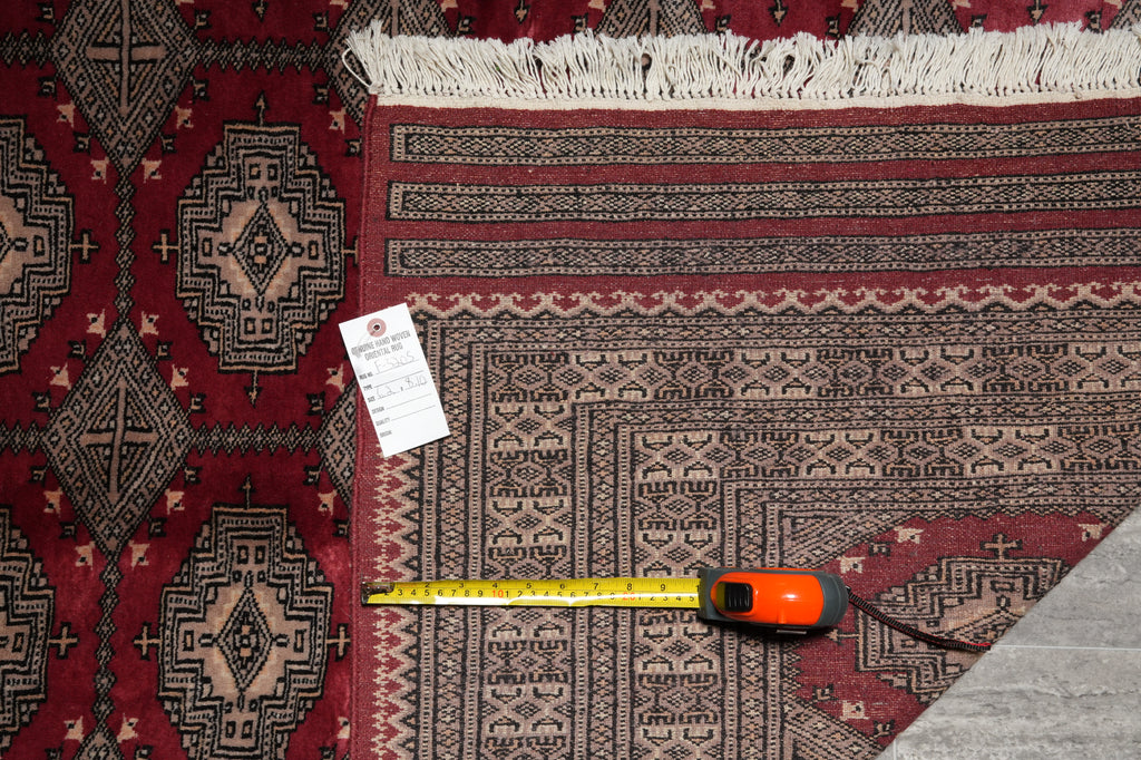 A close-up of a patterned area rug corner with fringe, a measuring tape, and a small tag placed on the rug to show scale. The rug has a deep red field with beige and black geometric motifs.