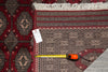 Load image into Gallery viewer, A close-up of a patterned area rug corner with fringe, a measuring tape, and a small tag placed on the rug to show scale. The rug has a deep red field with beige and black geometric motifs.