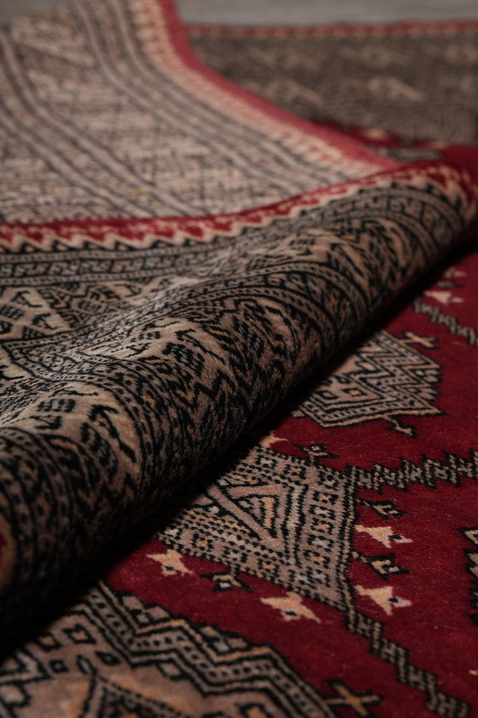 Close-up of a patterned rug with a folded section showing a deep red field, beige and black geometric motifs, and a dark central border.