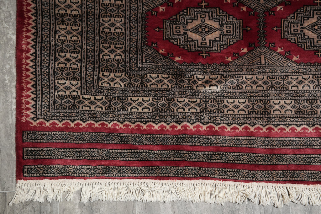 A close-up of a traditional patterned rug corner showing a red field, black and beige geometric motifs, decorative borders, and white fringe at the bottom.