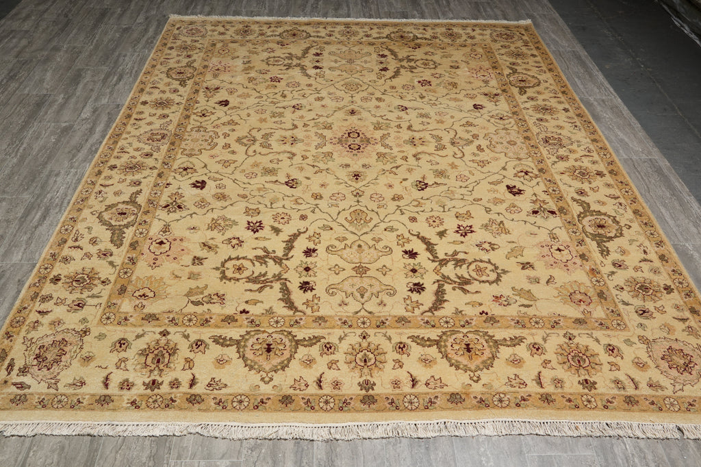 A rectangular area rug with an intricate traditional floral pattern, displayed on a gray wood-plank floor. The rug features a warm beige base with brown, gold, and muted pink accents in its detailed medallion and border motifs.