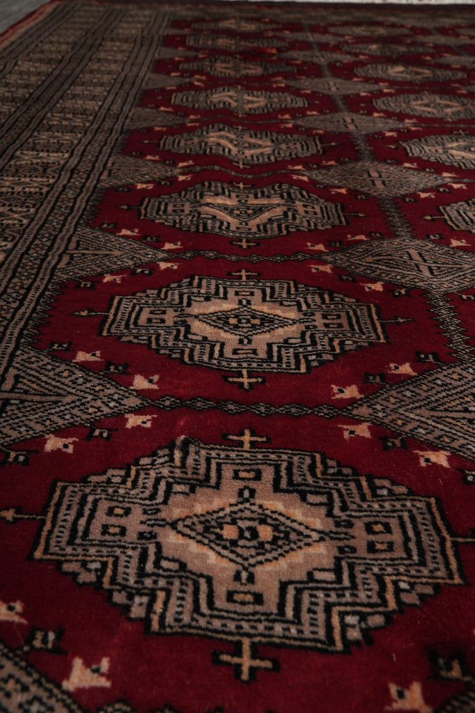 A close-up view of a traditional patterned rug with a deep red field, beige and black geometric medallions, and small cream accents.