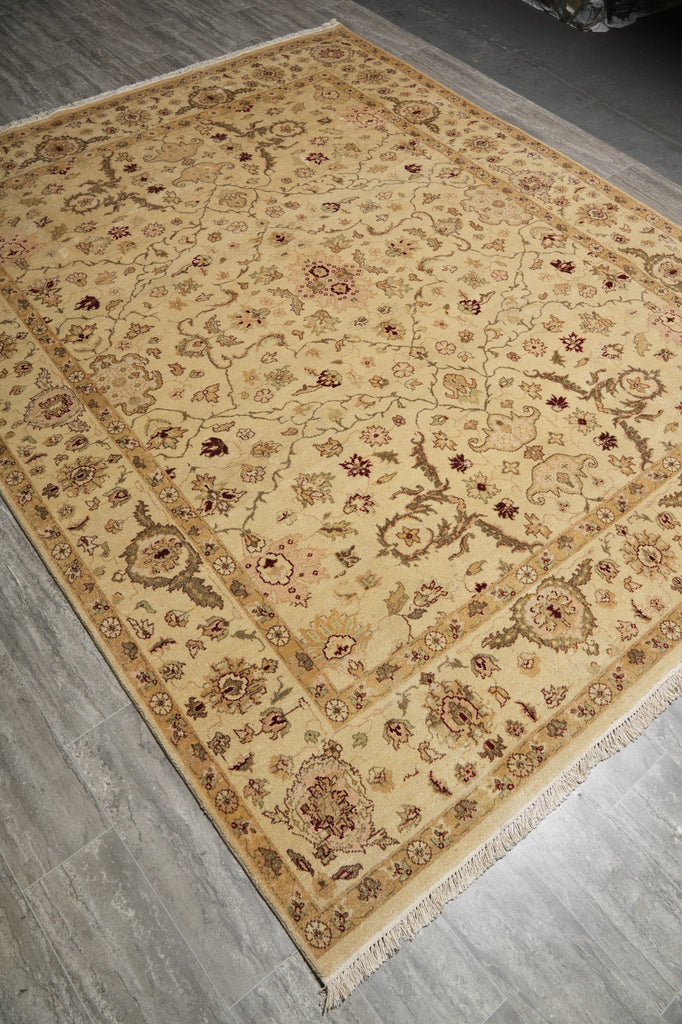 A rectangular area rug with an intricate traditional floral pattern, shown laid out on a gray wood-look floor. The rug has a warm beige base with brown, rust, and muted green details, and features a fringed edge.