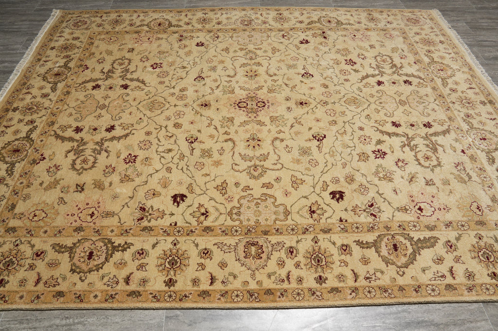A rectangular area rug with an intricate traditional floral and medallion pattern, laid out on a gray tiled floor. The rug features a warm beige base with brown, muted red, and gray accents in its detailed motifs and borders.
