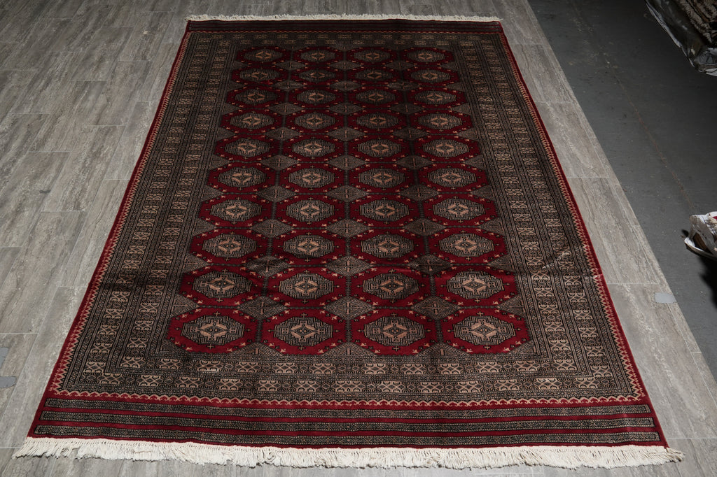 A rectangular area rug with a traditional geometric pattern in deep red, dark brown, and beige, featuring white fringe on the bottom, laid out on a gray wood-look floor.